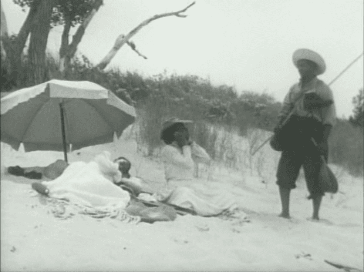 1. This film still is from the film Compensation, and is of three Black people on a beach. Two femmes in dresses rest on a blanket in long white dresses, one lays under a beach umbrella - maybe taking a nap. The other sits up and looks at a man who she is communicating with. Her hands cover her ears and the man stands next to her holding a ukulele, wearing a bag slung over his shoulder and a large harpoon pole for catching fish.