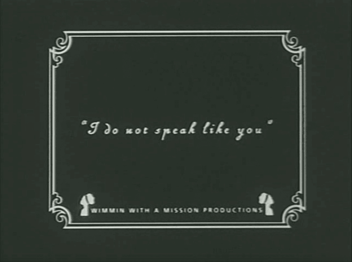 4. Another still from the same film, its a slide decorated with Art Deco edges and a design of two heads with the words between them “WIMMIN WITH A MISSION PRODUCTIONS”. In the middle of the designed slide is the text “I do not speak like you”. 