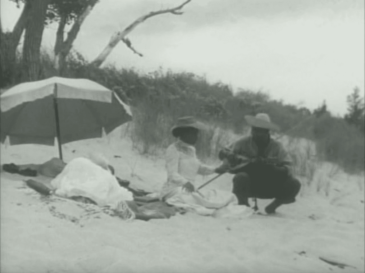 6. Back to the wide view of the three people on the beach we see the man squatting down next to the woman. Her hand is on the ukulele and he seems to be strumming it. We can infer that she is feeling the vibration of the strum. 