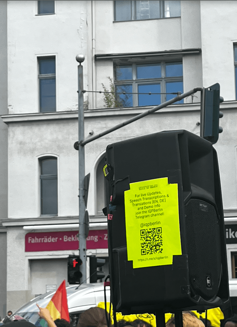 1. This is a photo of a big speaker with a neon paper taped to it. The tops of heads in the street are shown, no faces are visible. There peaks out a rainbow flag on the bottom left of the image. The neon paper has a big QR code on it, a URL and some text inviting protestors to join a telegram chat for live updates, transcriptions & translations of the demo info IQP Berlin. 