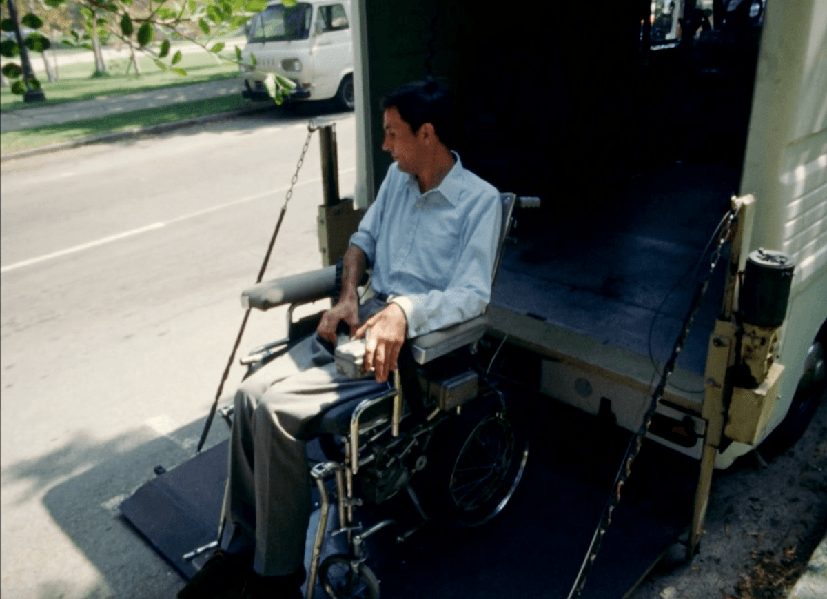 1. Shown is a screenshot from the film Behinderte Zukunft / Disabled Future which is of Adolf Ratzka on a lifting board moving into a box truck with himself & his wheelchair. Its a sunny day in the film and Ratzka appears as a white German physically disabled man sitting in a wheelchair - wearing a button down shirt and gray slacks. 