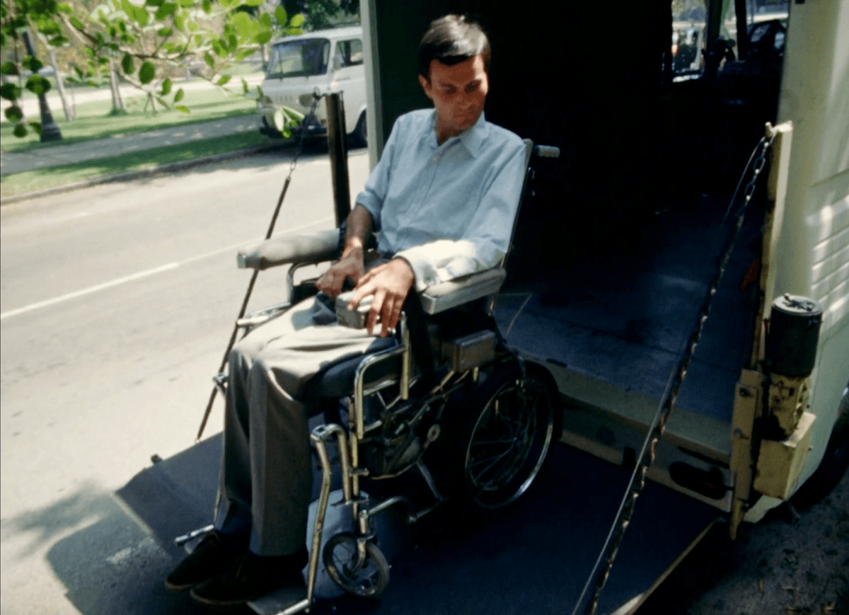 2. Shown is a screenshot from the film Behinderte Zukunft / Disabled Future which is of Adolf Ratzka on a lifting board moving into a box truck with himself & his wheelchair. Its a sunny day in the film and Ratzka appears as a white German physically disabled man sitting in a wheelchair - wearing a button down shirt and gray slacks. In this image Ratzka is looking down at the pavement as the board moves up. 