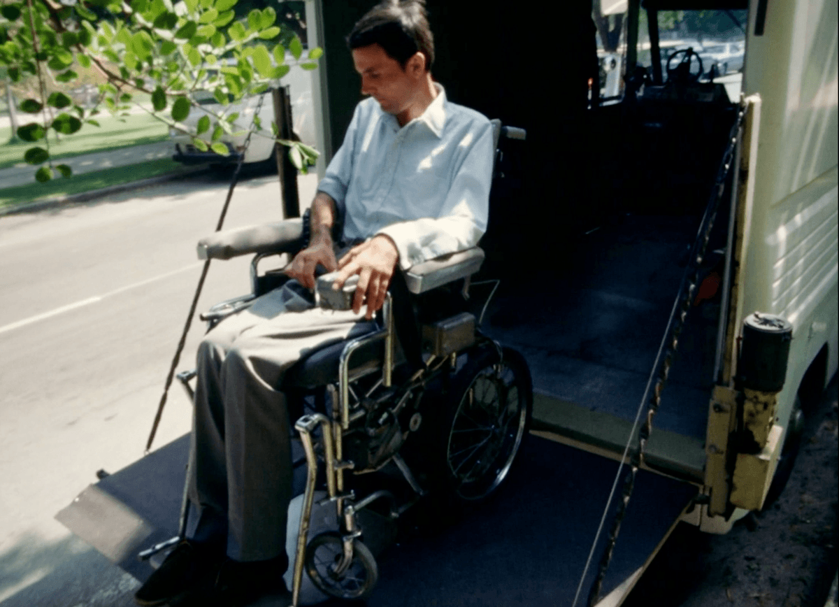 3. Shown is a screenshot from the film Behinderte Zukunft / Disabled Future which is of Adolf Ratzka on a lifting board moving into a box truck with himself & his wheelchair. Its a sunny day in the film and Ratzka appears as a white German physically disabled man sitting in a wheelchair - wearing a button down shirt and gray slacks. In this image Ratzka and his wheelchair has been lifted by the mechanized board nearly into the box truck. 