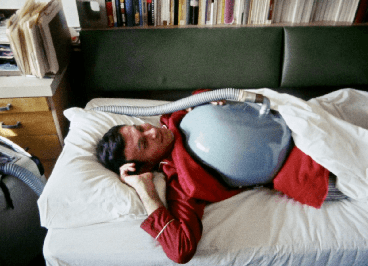 1. In this image Adolf Ratzka is laying in bed wearing red pajamas. Behind him are many books on a shelf. Over his chest he has a large blue iron half dome that has a tube connected to it. He rests on the pillow with a half smile on his face.